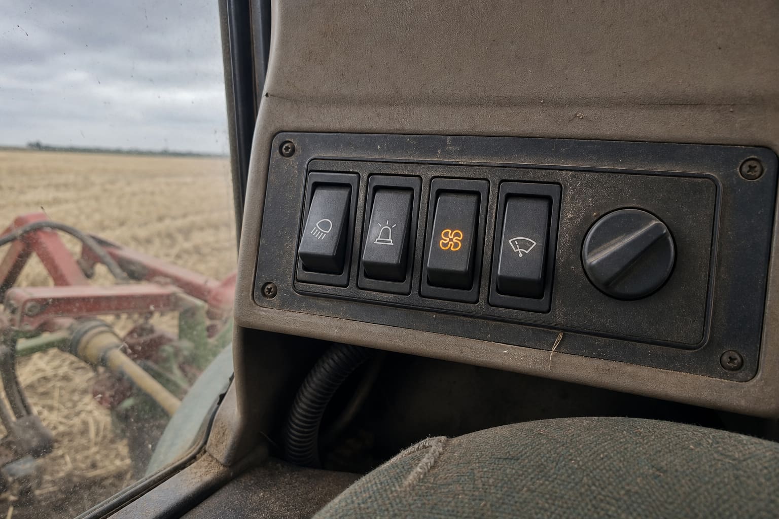 Sealed rocker switch panel and rotary control in a dusty agricultural machine cab, with a corrugated harness in the recess below and a harvested stubble field through the side window